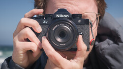 Photographer holding a Nikon Z4 mirrorless camera with a 50mm lens toward the camera against a blue sky and ocean backdrop.