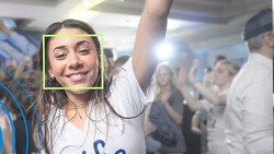 Smiling woman with arm raised at crowded indoor event, face detection box overlay visible.