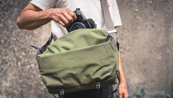 Photographer removing a DSLR camera from a sage green canvas messenger bag with white strap.