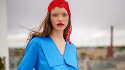 Woman wearing bright blue wrap blouse and red lace headwrap looking over shoulder outdoors.