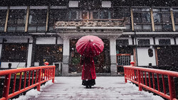 Person holding red umbrella walking across red bridge in heavy snowfall toward traditional architecture.