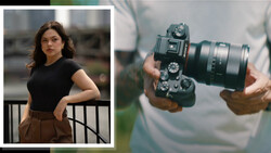 Portrait of a woman in black top and brown skirt standing against an urban railing; detail of hands holding a mirrorless camera with telephoto lens.