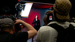 Audience members photograph a speaker on stage lit by professional studio lighting against a red and blue backdrop.
