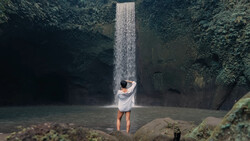 Person in white shirt standing in shallow water beneath a tall waterfall surrounded by lush vegetation.