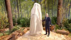 Man in dark suit unveiling a white draped sculpture in a forest setting with large trees and natural ground cover.