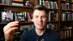Photographer holding a compact camera while smiling at the camera in front of a wooden bookshelf.