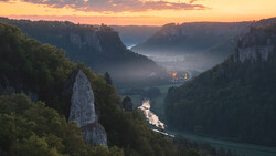 Dramatic river valley at sunrise with steep forested cliffs, mist, and a solitary rock formation in the foreground.