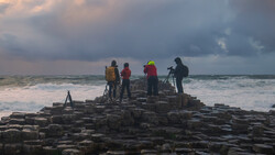 Four people with cameras and tripods standing on rocky coastal outcrop during overcast conditions.