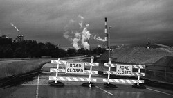 Road closed barricades blocking a highway with industrial smokestacks visible in the distance under dark skies.