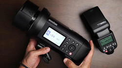 Hands holding a Godox studio flash with LCD screen and a Nikon TTL speedlight for comparison.