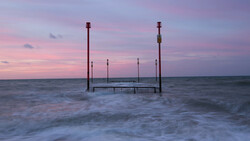 Wooden pier structure with tall posts standing in shallow ocean waves at dusk, with pink and purple sky.