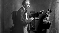 Man in suit gesturing expressively while holding a microphone in a studio setting.