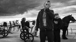 Man in dark coat and mask stands in foreground with horses and carts in dramatic black and white scene.