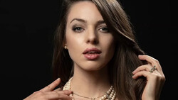 Woman with long dark hair wearing pearl necklace and rings against black background.