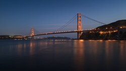 Suspension bridge illuminated at dusk, spanning a bay with calm water reflecting its lights.
