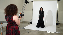 Photographer shooting a portrait of a woman in a black dress against a white backdrop in a studio setting.