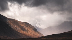 Dramatic mountain landscape with layered peaks under stormy clouds and golden light breaking through mist.