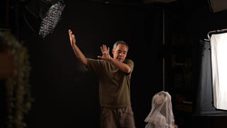 Director gesturing with hands raised during film production on a dark set with professional lighting equipment visible.
