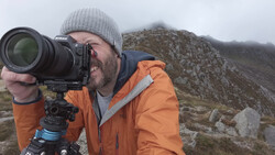 Photographer with Nikon camera on tripod looking through telephoto lens in mountainous terrain.