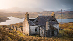 Dilapidated stone cottage on moorland with mountains and lochs in background at golden hour.