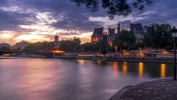 Illuminated riverside buildings at twilight reflected in still water under a dramatic purple and gold sky.