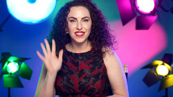 Woman with curly dark hair waving at camera against colorful studio lighting setup.