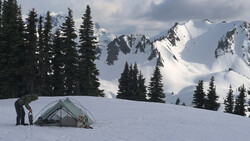 Backpacker standing beside a dome tent on snow-covered slope with snow-capped peaks and evergreen forest in background.