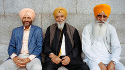 Three men with turbans seated against a stone wall, wearing formal jackets and traditional head coverings.