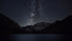 Milky Way galaxy stretches across a starlit night sky above snow-capped mountains and a dark alpine lake.