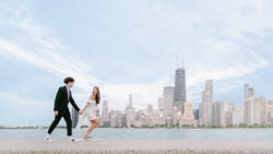 Couple in formal attire playfully running on a beach with a city skyline in the background.