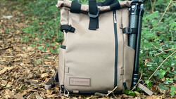 Tan canvas camera backpack with black straps and tripod attachment, photographed outdoors on forest ground.
