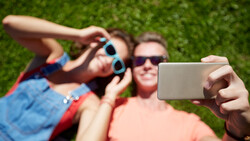 Three people taking a selfie outdoors on grass, with one person holding a smartphone in the foreground.