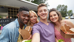 Four young adults smiling at a food truck window while holding takeout containers.