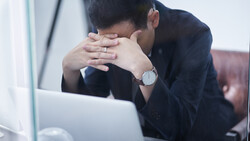 Man in dark blazer covering his face with both hands while seated at laptop, appearing stressed or fatigued.