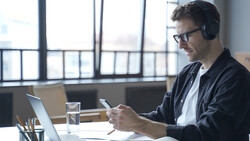 Man wearing headphones at desk working on laptop and smartphone in bright office with city view.