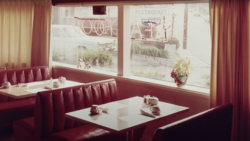 Retro diner booth with burgundy vinyl seating and white Formica table beside large storefront windows.