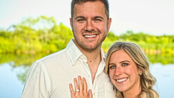 Smiling couple posing outdoors with water and green foliage in soft-focus background.