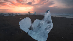 Illuminated icebergs on black sand beach at sunset with silhouetted figures in background.