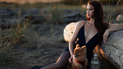 Woman in dark swimsuit seated on driftwood at beach during golden hour, holding small dog.
