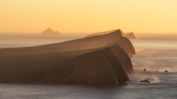 Dramatic coastal cliffs silhouetted against a golden sunrise over misty ocean waters.