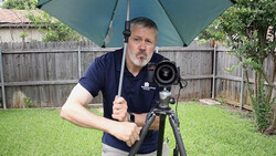 Videographer holding camera on tripod under umbrella in backyard setting.