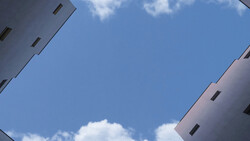Looking upward between two white apartment buildings framing a blue sky with white clouds.