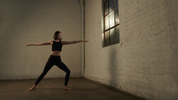 Woman in warrior pose III yoga position inside industrial brick studio with natural window light.