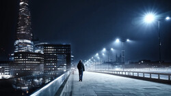 Solitary figure walking across an illuminated bridge at night with a modern city skyline in the background.