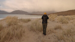 Runner in yellow hat jogging through sandy beach with dune grass toward misty mountains and water.