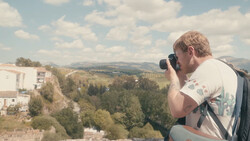Photographer holding camera to face, composing a shot of rolling hills and forested landscape.