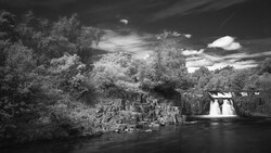 Black and white landscape with waterfall, river, and dense trees under dramatic cloudy sky.