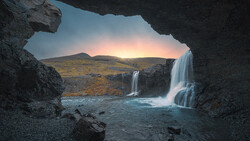 Waterfall cascading into turquoise pool, framed by dark rock cave opening with mountain landscape and sunset sky beyond.