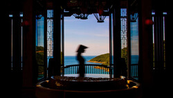 A blurred figure moves through an ornate pavilion overlooking a coastal landscape at twilight.