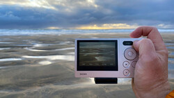 Hand holding a compact digital camera displaying a coastal landscape on its rear screen.
